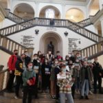 Uczestnicy w maseczkach stają na schodach holu budynku głównego AGH. W tle godło akademii i posąg S. Staszica.