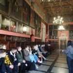 Uczniowie siedzą w auli Jagiellońskiej i słuchają przewodniczki. Na ścianach widoczne portrety postaci związanych z Uniwersytetem Jagiellońskim.