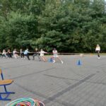 Grupa młodzieży bierze udział w zabawie przeciąganie linii, przed nimi leża kolorowe kółka hula-hop, krzesła, w tle widać zielone liście na drzewach