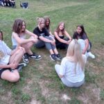 Grupka dziewcząt siedzi na zielonej trawie blisko siebie w półokręgu, uśmiechają się, w tle inna grupa siedzi obok siebie