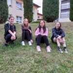 Cztery dziewczyny siedzą na trawie, w tle tuje, budynek szkoły i budynek hali sportowej
