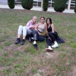 Trzy dziewczyny siedzą blisko siebie na trawie, uśmiechają się, w tle budynek hali sportowej