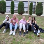 Grupa pięciu dziewczyn siedzi na trawie, uśmiechają się, w tle budynek hali sportowej i zielone tuje