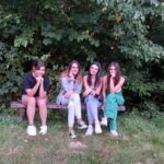 Grupa czterech dziewczyn siedzi na drewnianej ławce, uśmiechają się, w tle zielona trawa i zielone liście na drzewach