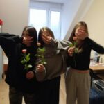 Trzy dziewczyny stoją, zakrywają dłońmi twarze, trzymają róże w rękach.